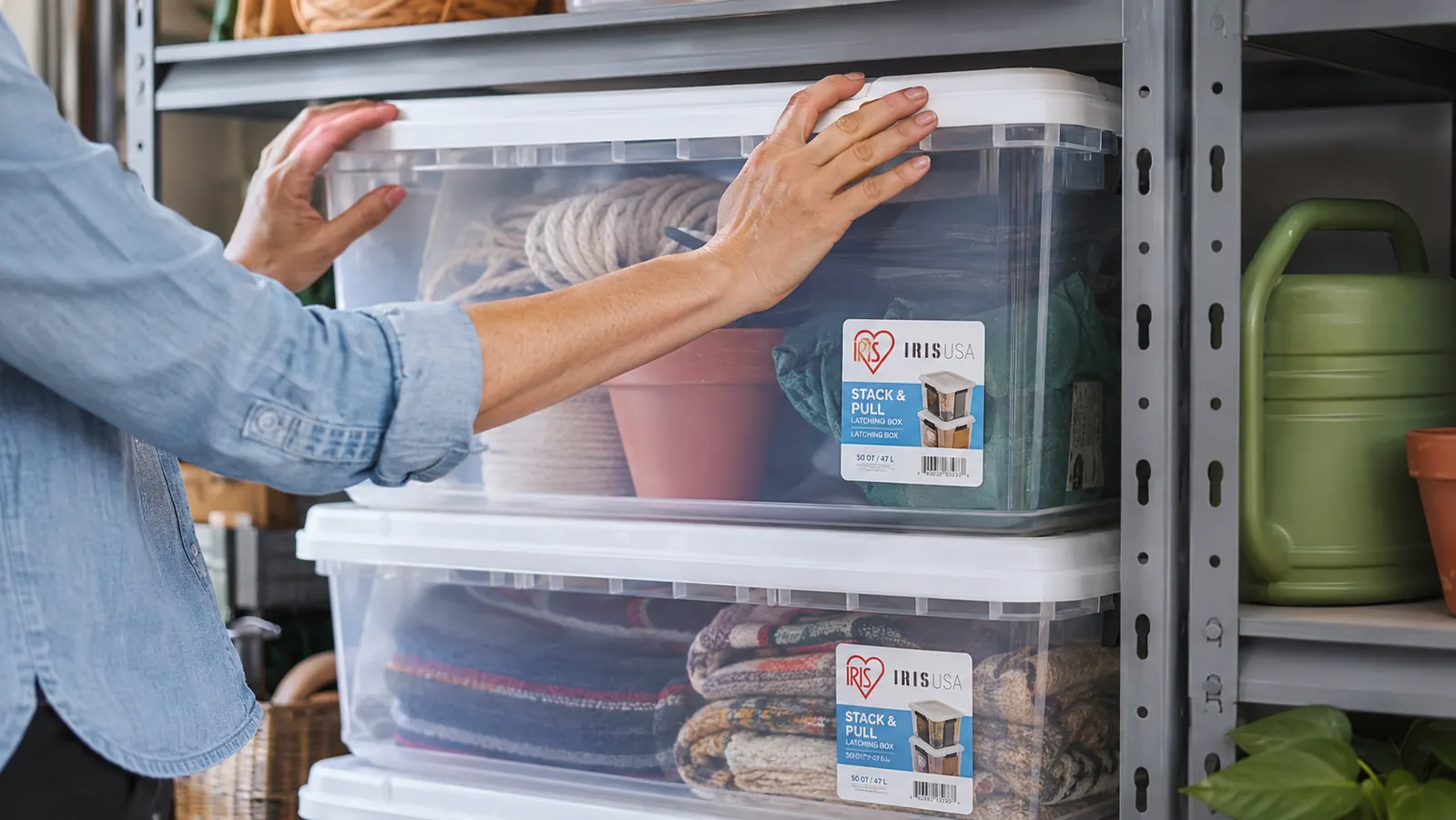 Hands placing a lidded clear storage bin onto a stack of two identical bins on metal garage shelving